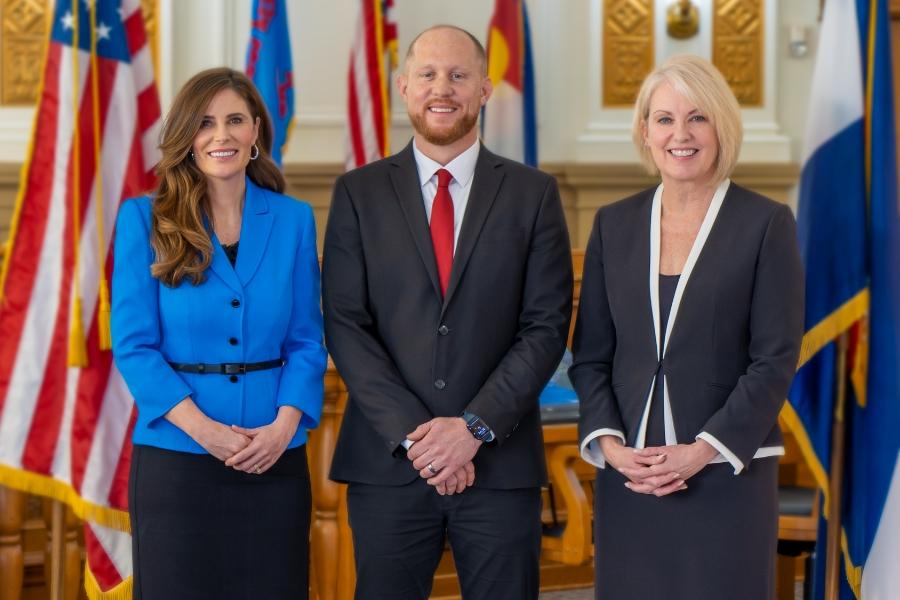 Three individuals standing next to each other smiling, in professional clothes. One man in the middle and two women beside him on each side. 
